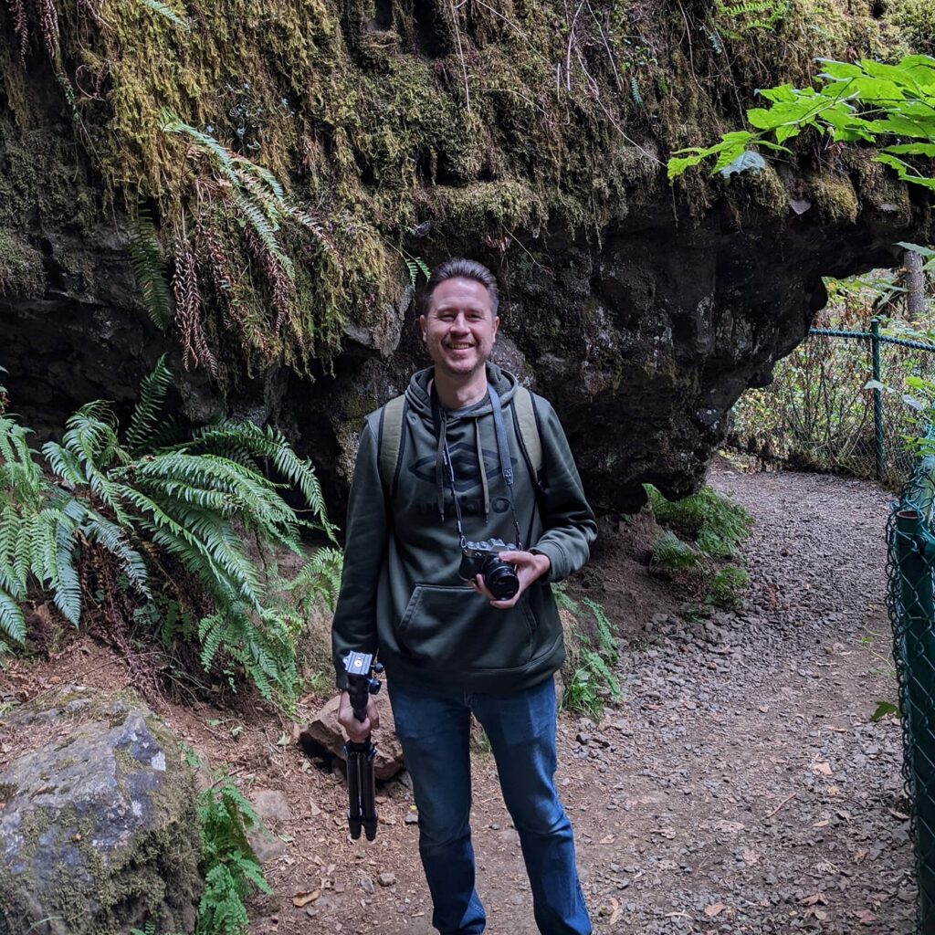 Headshot of Kyle McPherson smiling with camera equipment in front of landscape greenery.