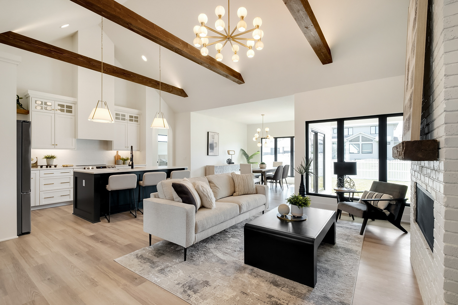 A white and grey toned, open concept living room with high ceilings and dark wooden support beams and windows looking into the backyard.